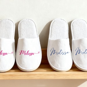 以下が含まれることがあります： ピンクと青の筆記体で「Melissa」という名前がプリントされた白いスパ用スリッパが4足。 スリッパは木製の棚に並べられています。 スリッパは柔らかく、豪華な素材で作られています。