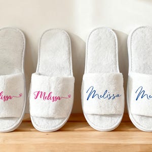Può includere: Quattro pantofole da spa bianche in peluche con il nome "Melissa" stampato sopra. Due pantofole hanno il nome in rosa e due in blu. Le pantofole sono esposte su una superficie di legno.