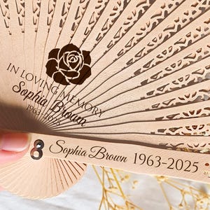 Op de afbeelding: Een houten handwaaier met een delicaat kantpatroon. De waaier is gesloten en heeft een zwarte roos en de tekst "In loving memory Sophia Brown 1963-2025" gegraveerd op de voorkant.