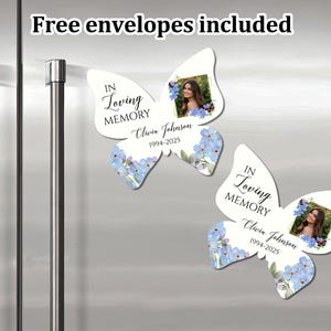 K&ouml;nnte beinhalten: Zwei wei&szlig;e, schmetterlingsf&ouml;rmige Erinnerungsmagnete mit blauen Vergissmeinnicht-Blumen. Jeder Magnet hat einen Fotorahmen und den Text "In Loving Memory" mit einem Namen und Daten. "Free envelopes included" steht oben im Bild.