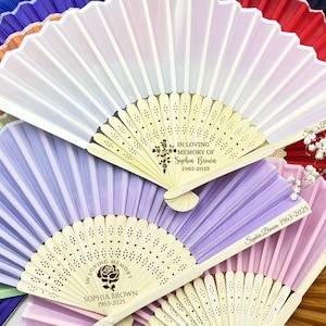 May include: A set of eight hand fans with wooden handles and white silk fabric. The fans are decorated with a floral design and the text "In loving memory of Sophia Brown 1963-2025".