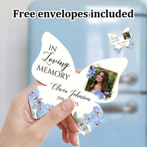 K&ouml;nnte beinhalten: Wei&szlig;e, schmetterlingsf&ouml;rmige Gedenkkarte mit einem Foto einer Frau und dem Text "In Loving Memory Olivia Johnson 199_ _25". Die Karte ist mit blauen Vergissmeinnicht-Blumen auf wei&szlig;em Hintergrund verziert.