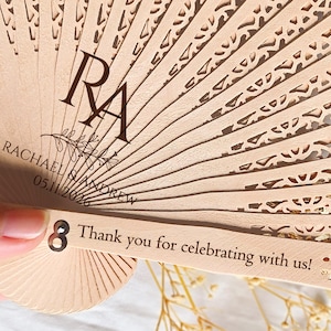 May include: A wooden hand fan with a decorative cut-out design. The fan is personalized with the names "Rachael & Andrew" and the date "05.11.2026". The fan also has the initials "RA" and the text "Thank you for celebrating with us!"
