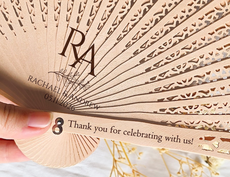 May include: A wooden hand fan with a decorative cut-out design. The fan is personalized with the names "Rachael & Andrew" and the date "05.11.2026". The fan also has the initials "RA" and the text "Thank you for celebrating with us!"