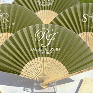 May include: Olive green personalized wedding fans with white text. The fans display initials, names, and dates: Sophia & Robert 09.14.2028, Rachel & Justin 09.14.2028, and S R 09.14.2028. The fans have a wooden base and are displayed on a white surface.