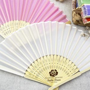 May include: Two white and pink hand fans with wooden handles. The white fan has a floral design and the text "In loving memory Sophia Brown 1963-2023" printed on the handle. The pink fan is plain.