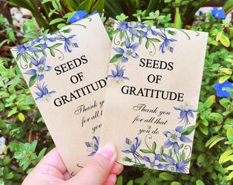 Może przedstawiać: Dwa brązowe papierowe opakowania nasion z ilustracjami niebieskich i białych kwiatów. Tekst na opakowaniach brzmi "Seeds of Gratitude" z wiadomością z podziękowaniem.