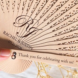 May include: A wooden hand fan with a white and gold design. The fan has the text "Rachel & Justin 05.21.2026" and "Thank you for celebrating with us!" printed on it.