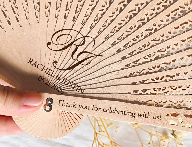 May include: A wooden hand fan with a white and gold design. The fan has the text "Rachel & Justin 05.21.2026" and "Thank you for celebrating with us!" printed on it.