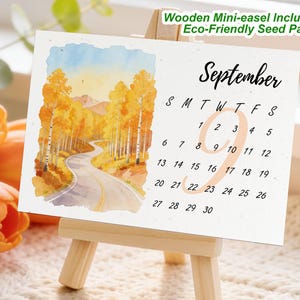 May include: A September calendar on eco-friendly seed paper, displayed on a wooden mini-easel. The calendar features a watercolor painting of a winding road through a forest of yellow trees. The text "September" is written in a script font.