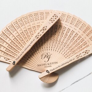 May include: Wooden wedding fan with a decorative cut-out design. The fan is personalized with the couple's names, "Rachel & Justin", and their wedding date, "05.21.2026". The text "Thank you for celebrating with us!" is engraved on both sides of the fan.