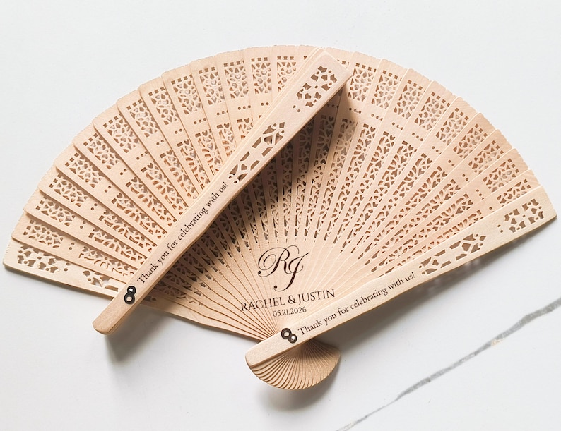 May include: Wooden wedding fan with a decorative cut-out design. The fan is personalized with the couple's names, "Rachel & Justin", and their wedding date, "05.21.2026". The text "Thank you for celebrating with us!" is engraved on both sides of the fan.