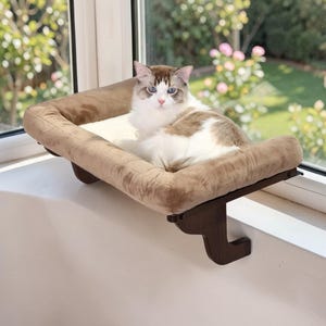 May include: A brown cat window perch with a plush, beige cushion. The cat bed is mounted on a dark brown wooden frame and is attached to a window sill. A fluffy, white and brown cat is resting on the bed, looking at the camera.
