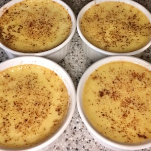 May include: Four ramekins filled with a creamy, light yellow custard, each topped with a sprinkle of brown spice. The ramekins are white and round, arranged in a square formation. The dessert appears freshly made and ready to serve.