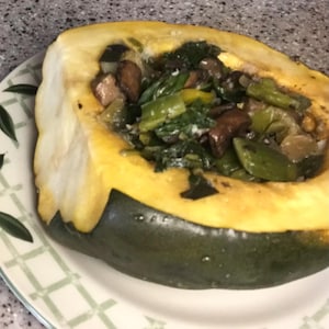 May include: A roasted acorn squash half with a dark green skin and yellow flesh, filled with a cooked mixture of dark green leafy vegetables, brown mushroom slices, and small pieces of yellow and green peppers, served on a patterned plate.