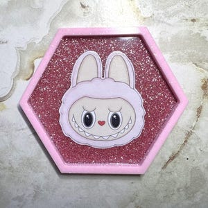 May include: A pink hexagonal resin tray with a glittery interior. The tray features a cartoon bunny character with large eyes, a small red heart, and a toothy grin. The bunny is wearing a pink hood.