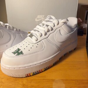 Może przedstawiać: Białe buty sportowe Nike Air Force 1 z zielono-białym logo z boku. Podeszwa ma tęczowy pasek. Logo Nike jest widoczne z boku buta. Buty stoją na drewnianej powierzchni.