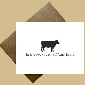 Puede incluir: Tarjeta de felicitación blanca con una silueta negra de una vaca y el texto "holy cow, you're halfway home". La tarjeta está dentro de un sobre marrón, sobre un fondo beige.