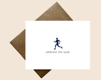 May include: A white rectangular greeting card featuring a dark blue silhouette of a person running. Below the runner, the text "embrace the suck" is printed in a dark blue typewriter-style font. A brown envelope is positioned behind the card.