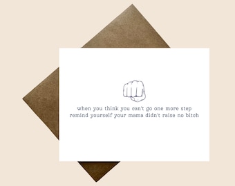 May include: A white greeting card featuring a dark blue line drawing of a fist and the text: when you think you can't go one more step remind yourself your mama didn't raise no bitch. The card is positioned against a brown envelope.