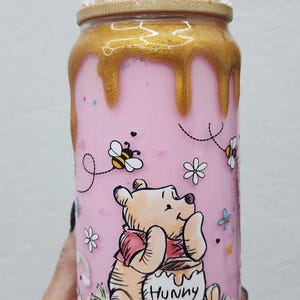 May include: Pink tumbler featuring Winnie the Pooh with a honey pot, bees, and flowers. The tumbler has a gold-colored lid and a clear straw. The top is decorated with whipped cream and sprinkles.