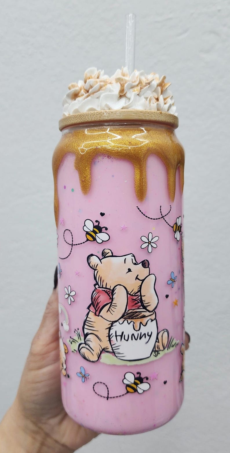 May include: Pink tumbler featuring Winnie the Pooh with a honey pot, bees, and flowers. The tumbler has a gold-colored lid and a clear straw. The top is decorated with whipped cream and sprinkles.