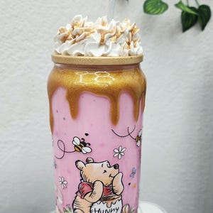 May include: A pink tumbler with a gold rim and dripping design, featuring a Winnie the Pooh illustration. The tumbler is topped with whipped cream and a straw. The design includes bees, flowers, and the word "Hunny".