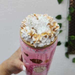 May include: Pink tumbler with a decorative design, topped with a swirl of white whipped cream and caramel drizzle. The tumbler has a wooden lid and is decorated with cartoon images. A refreshing beverage.