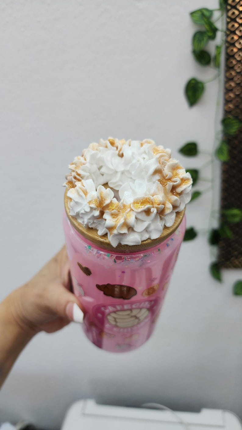 May include: Pink tumbler with a decorative design, topped with a swirl of white whipped cream and caramel drizzle. The tumbler has a wooden lid and is decorated with cartoon images. A refreshing beverage.