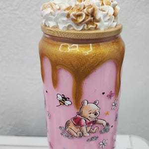 May include: A pink tumbler featuring Winnie the Pooh illustrations, a golden honey-like design, and a whipped cream topping. The tumbler has a wooden lid and a clear straw. The design includes bees and flowers.