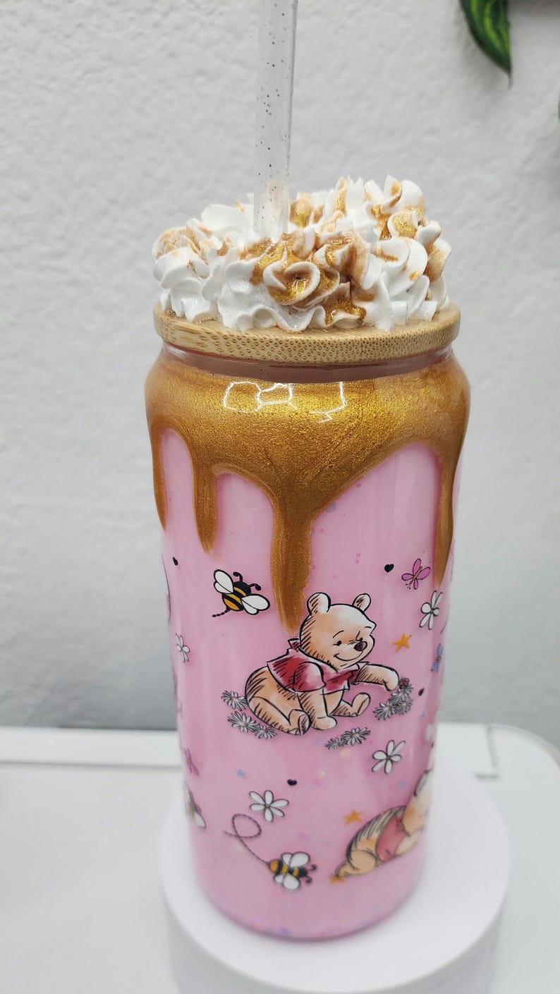 May include: A pink tumbler featuring Winnie the Pooh illustrations, a golden honey-like design, and a whipped cream topping. The tumbler has a wooden lid and a clear straw. The design includes bees and flowers.