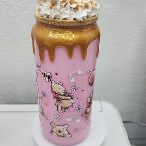 May include: A pink tumbler featuring Winnie the Pooh characters and floral designs. The top is gold with a dripping effect, topped with whipped cream. The tumbler is approximately 7 inches tall.