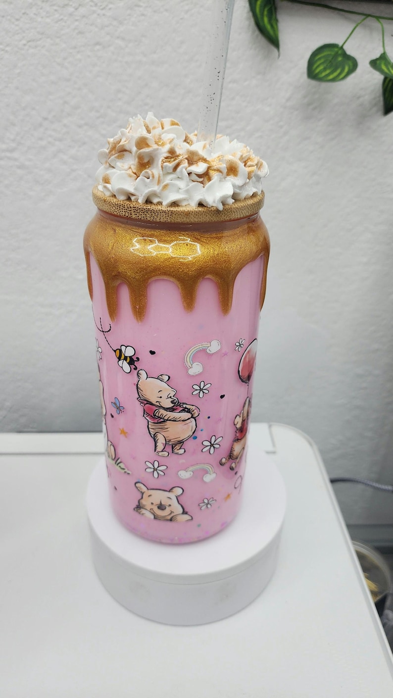 May include: A pink tumbler featuring Winnie the Pooh characters and floral designs. The top is gold with a dripping effect, topped with whipped cream. The tumbler is approximately 7 inches tall.