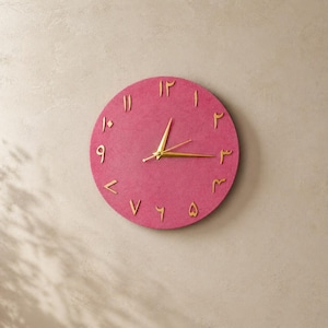 Könnte beinhalten: Eine runde, pinkfarbene Wanduhr mit goldfarbenen arabischen Ziffern und Zeigern. Das Zifferblatt hat eine strukturierte Oberfläche, und die Zeiger zeigen auf etwa 2:15 Uhr. Die Uhr ist ein dekorativer Artikel.