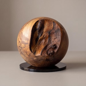 Könnte beinhalten: Eine kugelförmige Holzskulptur mit natürlicher Maserung in hellen und dunklen Brauntönen. Die Oberfläche weist eine tiefe, unregelmäßige Einkerbung auf, die die rohe Holzstruktur freilegt. Die Kugel ruht auf einem runden schwarzen Sockel.