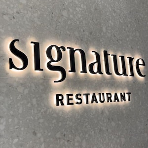 May include: A wall-mounted sign featuring the word Signature in a large, black, serif font above the word RESTAURANT in a smaller, black, sans-serif font. Both words are backlit with a warm white glow against a light gray speckled stone wall.