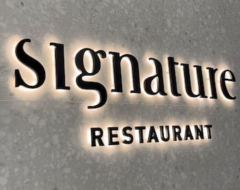 May include: A backlit sign for a restaurant. The word "Signature" is in a bold, black font above the word "Restaurant". The sign is mounted on a textured, light gray wall.