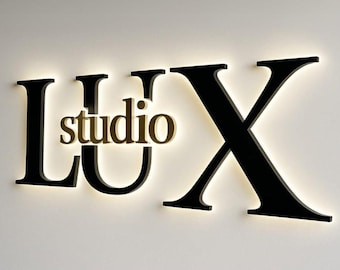 May include: Black "LUX" letters with a gold "studio" in the middle, all mounted on a white wall. The letters are illuminated from behind, creating a glowing effect. The design is modern and minimalist.