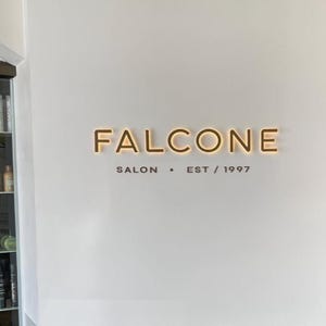 May include: A wall-mounted sign with the name "FALCONE" in illuminated, warm-toned letters. Below, the text "SALON &bull; EST / 1997" is printed in a dark, sans-serif font. The sign is centered on a white wall.