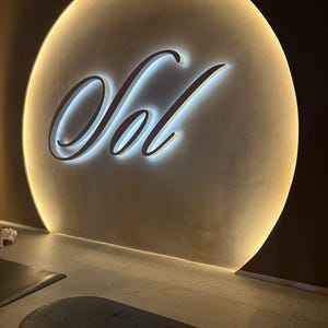 May include: A large, circular, beige wall feature with a backlit edge and the word Sol written in large, white, cursive, illuminated letters. Below the wall feature, a dark gray, rectangular exercise mat with the brand name Bala is visible.