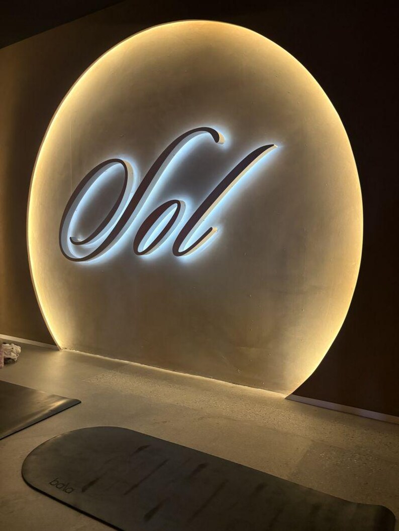 May include: A large, circular, beige wall feature with a backlit edge and the word Sol written in large, white, cursive, illuminated letters. Below the wall feature, a dark gray, rectangular exercise mat with the brand name Bala is visible.