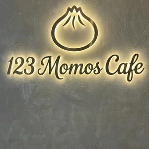 May include: A wall-mounted sign for 123 Momos Cafe featuring a backlit, dark-colored dumpling icon and the text 123 Momos Cafe in a cursive font. The sign is mounted on a textured, gray wall, creating a glowing halo effect around the lettering.