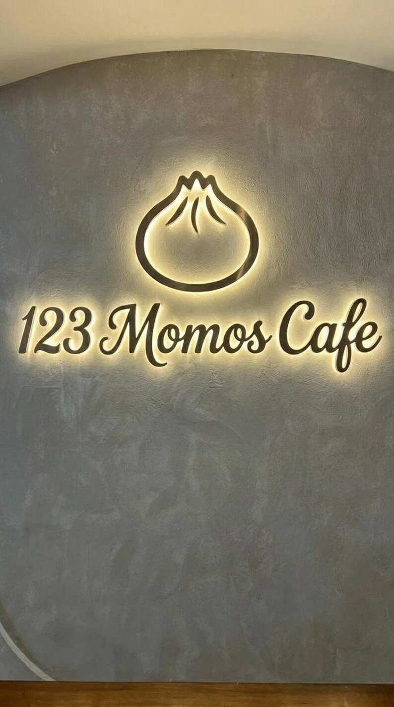 May include: A wall-mounted sign for 123 Momos Cafe featuring a backlit, dark-colored dumpling icon and the text 123 Momos Cafe in a cursive font. The sign is mounted on a textured, gray wall, creating a glowing halo effect around the lettering.