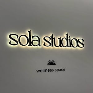 May include: Black illuminated sign that reads "sola studios" in a stylized font. Below the text is a graphic of a sunburst and the words "wellness space" in a sans-serif font.