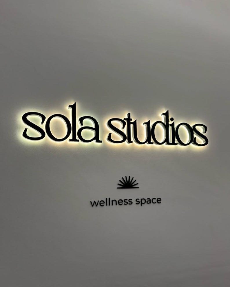 May include: Black illuminated sign that reads "sola studios" in a stylized font. Below the text is a graphic of a sunburst and the words "wellness space" in a sans-serif font.