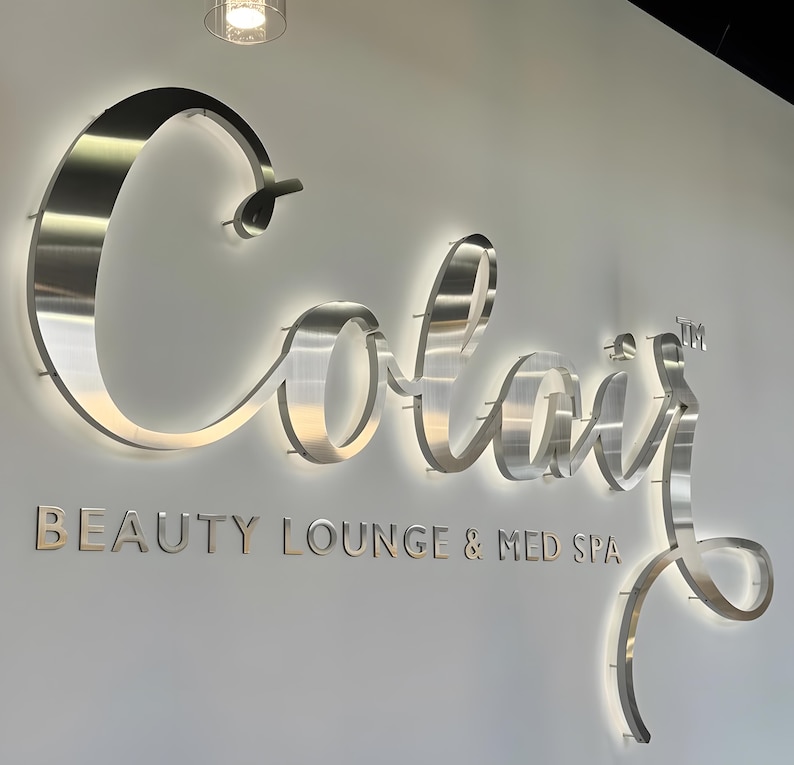 May include: A large, brushed metal wall sign featuring the word Colair in a cursive script font with a trademark symbol, backlit with a soft white glow. Below the main text, the words BEAUTY LOUNGE & MED SPA appear in a clean, sans-serif font.