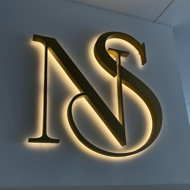May include: A gold-colored illuminated sign displaying the letters "NS" mounted on a white wall. The letters have a serif font and are lit from behind, creating a warm glow around the edges. The sign is a decorative element.