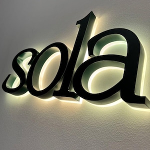 May include: Black, illuminated "sola" sign mounted on a light-colored wall. The letters are in a bold, elegant font, with a warm, glowing light emanating from behind them, creating a striking visual effect. The sign is a decorative piece.