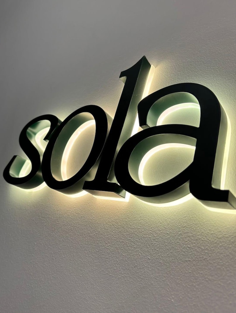 May include: Black, illuminated "sola" sign mounted on a light-colored wall. The letters are in a bold, elegant font, with a warm, glowing light emanating from behind them, creating a striking visual effect. The sign is a decorative piece.