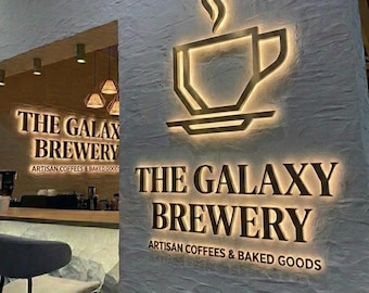 May include: A wall sign for The Galaxy Brewery featuring a backlit coffee cup icon with steam, the text The Galaxy Brewery, and the words Artisan Coffees & Baked Goods in dark lettering against a textured white wall with warm lighting.